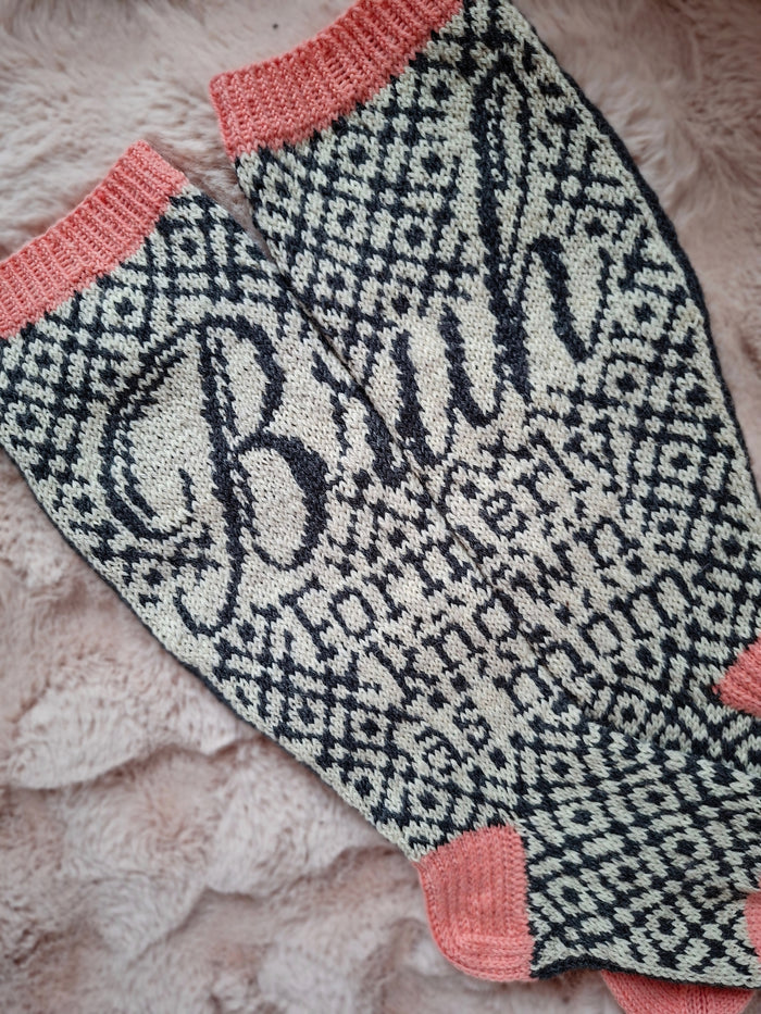 Knitted pair of socks with black and white pattern and pink trim and the text "Bruh Formerly known as mom" on a soft surface