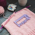 Pink knitted fabric with a ruler on top, yarn balls, and knitting tools in the background.