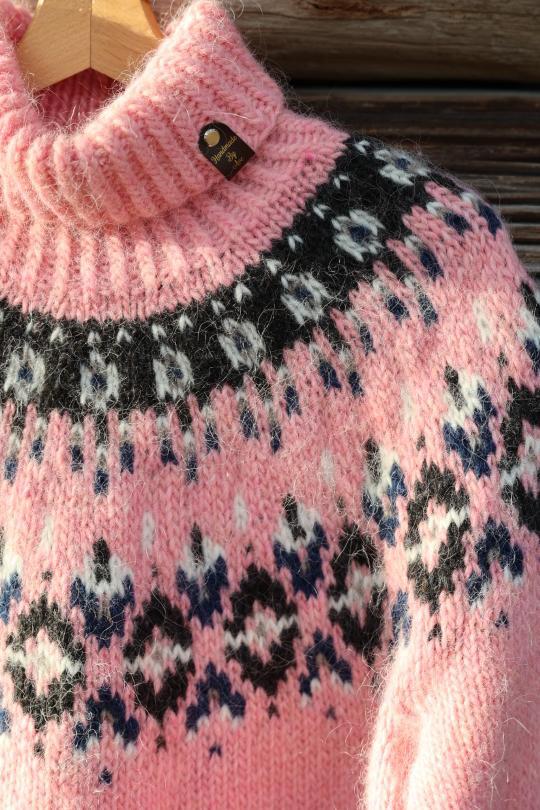 A close-up of a pink knit sweater with a black, white, and blue stranded colorwork circular yoke pattern, hanging on a wooden hanger against a rustic wooden background.
