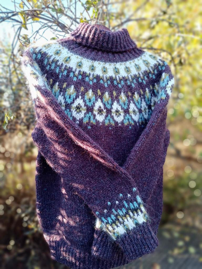 A hand-knit turtleneck sweater in a deep plum purple, featuring a light blue, white, and yellow stranded colorwork circular yoke pattern and matching patterned cuffs, photographed outdoors against a blurred background of green foliage.